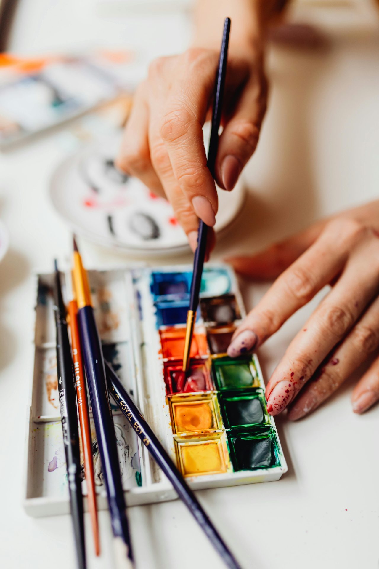 Hands blending vibrant watercolors on a palette with brushes, showcasing artistic creativity.